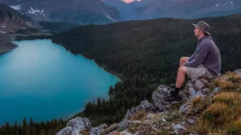 A person wearing a hat, gray hoodie, and shorts sits on a rocky ledge overlooking a blue lake surrounded by dense forest and mountains. The sun is setting in the background, casting a warm glow across the sky.