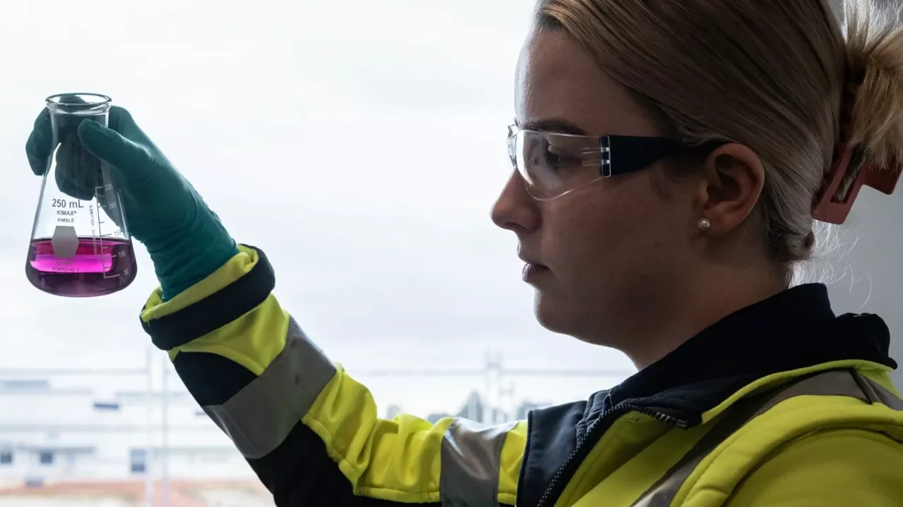 Woman in protective clothing and goggles examining liquid in lab beaker.