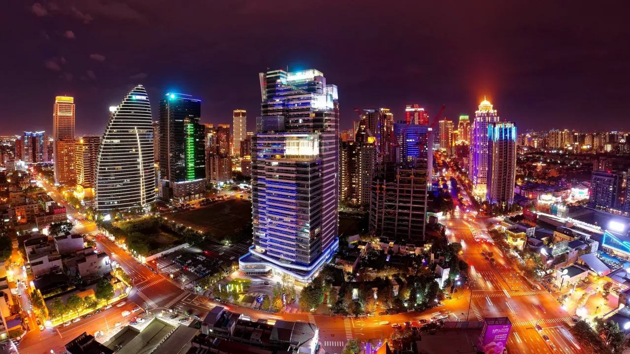 A panoramic nighttime cityscape showing illuminated skyscrapers and busy streets with glowing traffic lights and urban lights, showcasing a bustling metropolitan area.
