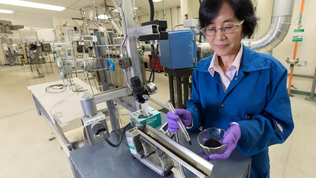 A female scientist in a lab coat conducts an experiment in an industrial setting, interacting with equipment while examining a sample in a mixing dish.