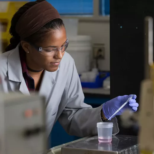 An Albemarle scientist wearing protective eyewear and gloves uses a pipette to add liquid to a small container in a laboratory. The scientist is focused on the task, and various lab equipment is visible in the background.